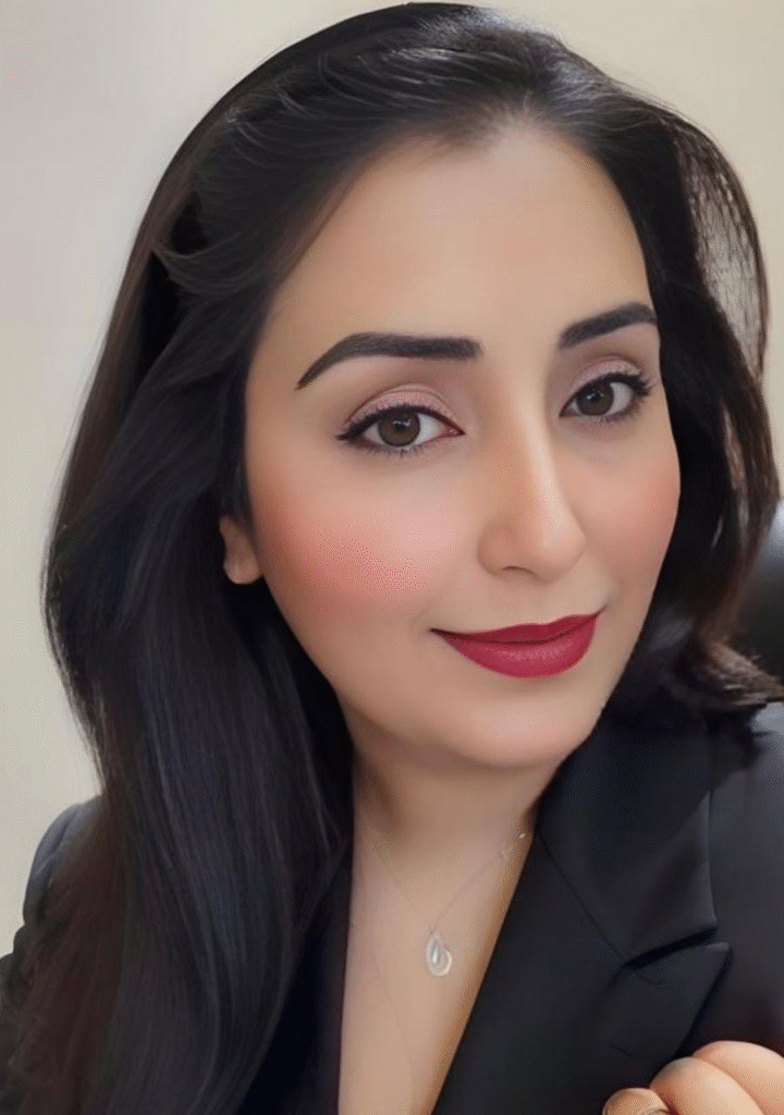 "Close-up of a woman with long black hair, wearing a black blazer, showcasing subtle makeup and a confident smile, with a delicate necklace visible in the shot."