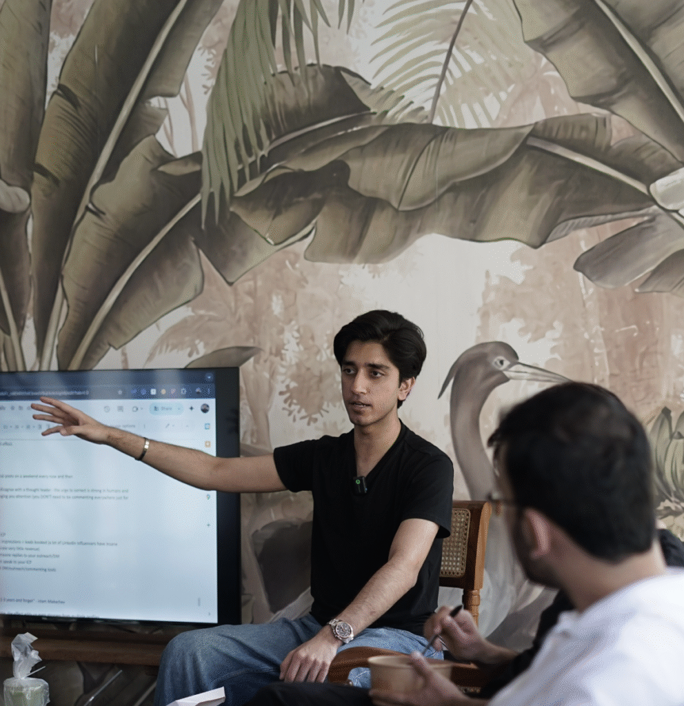 "Young professional giving a presentation to a group during a business meeting, with a large screen showing content behind them. The photo features a modern office setting with a nature-themed wall mural."