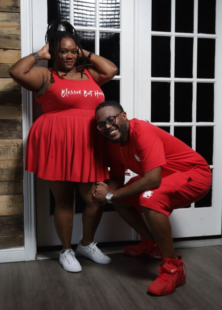 A couple dressed in matching red outfits, with the woman wearing a red dress that says "Blessed But Humble" and the man kneeling beside her, smiling, and dressed in a red T-shirt and shorts.