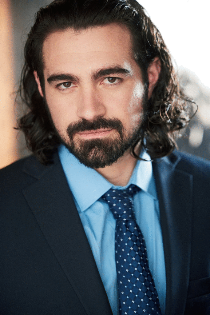 "Professional headshot of a man with long dark hair and a beard, wearing a blue suit and tie, looking directly at the camera with a serious expression."
