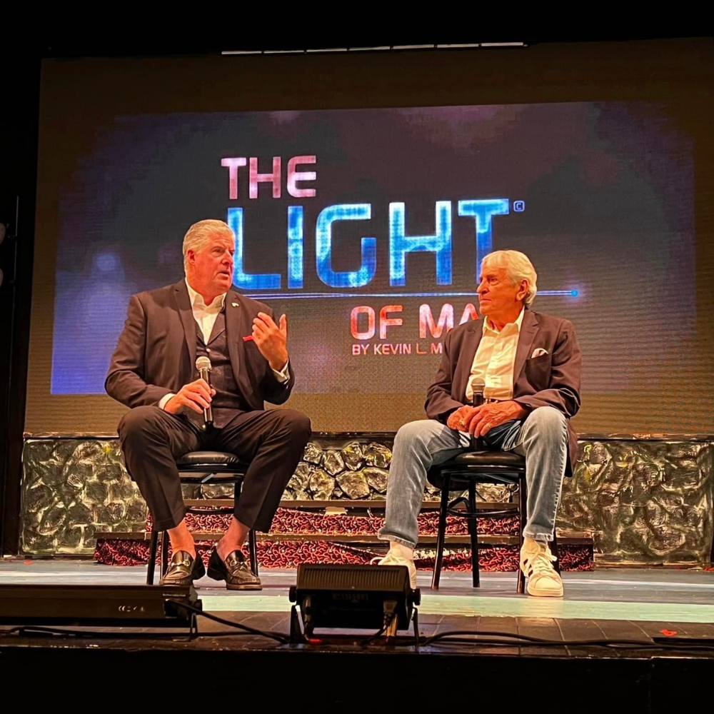 Two men sit on stage during a conversation about The Light of Man by Kevin L. McMullen, with a backdrop featuring the book's title and vibrant lighting.