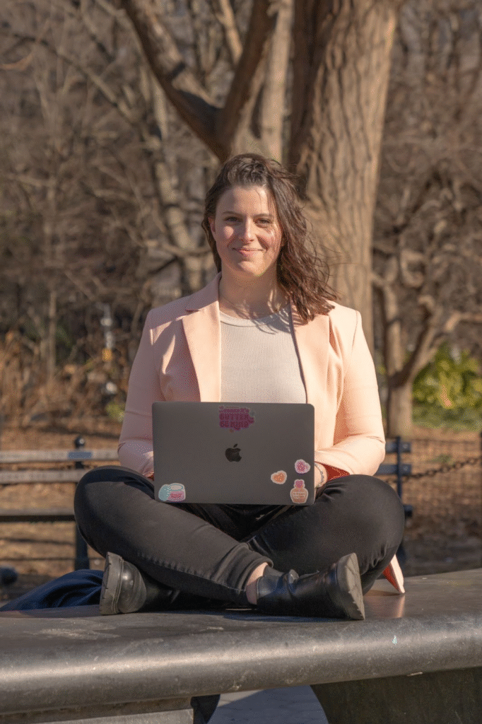 "A woman sitting outdoors at a park bench, working on her laptop while wearing a light pink blazer and black pants, with stickers on the laptop, creating a casual and productive vibe."