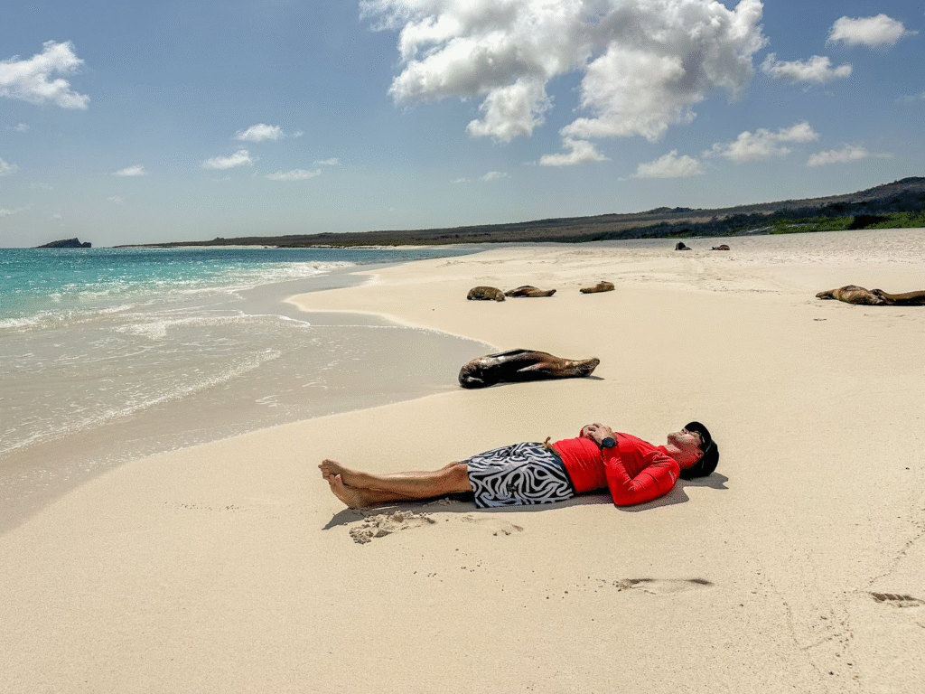Man relaxing on a pristine sandy beach in the Galápagos Islands, lying beside sea lions with turquoise ocean waves and a scenic horizon in the background.