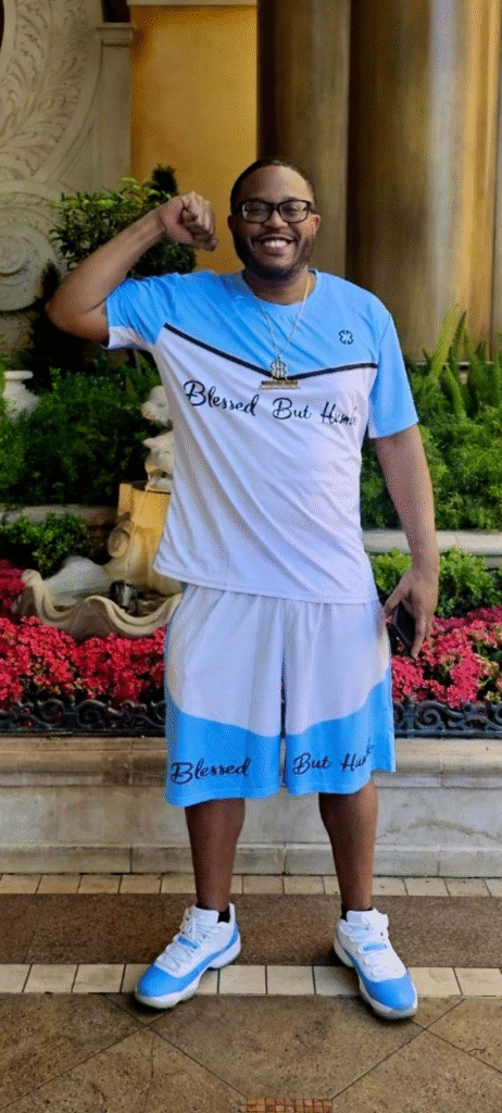 A man flexing his arm confidently, wearing a blue and white sports outfit with the phrase "Blessed But Humble" on it, posing outdoors in front of vibrant green plants and flowers.