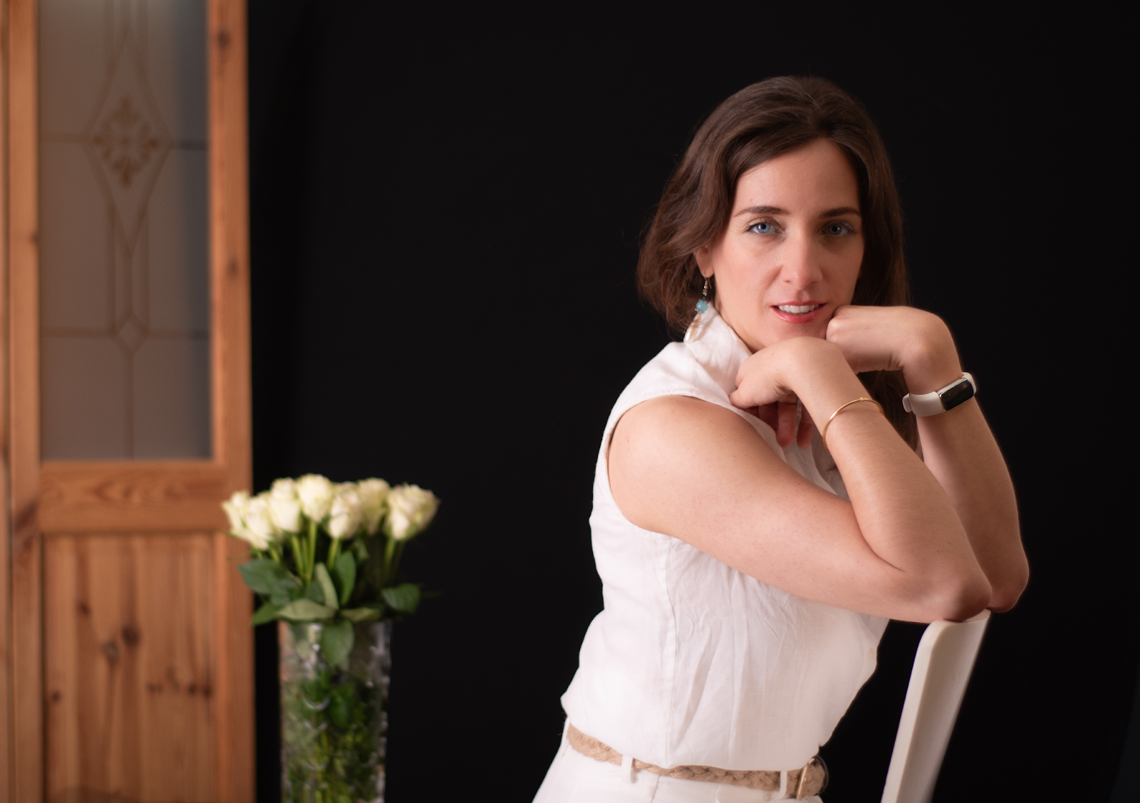 Portrait of a woman dressed in a white sleeveless top, posing confidently with a bouquet of white roses in the background.