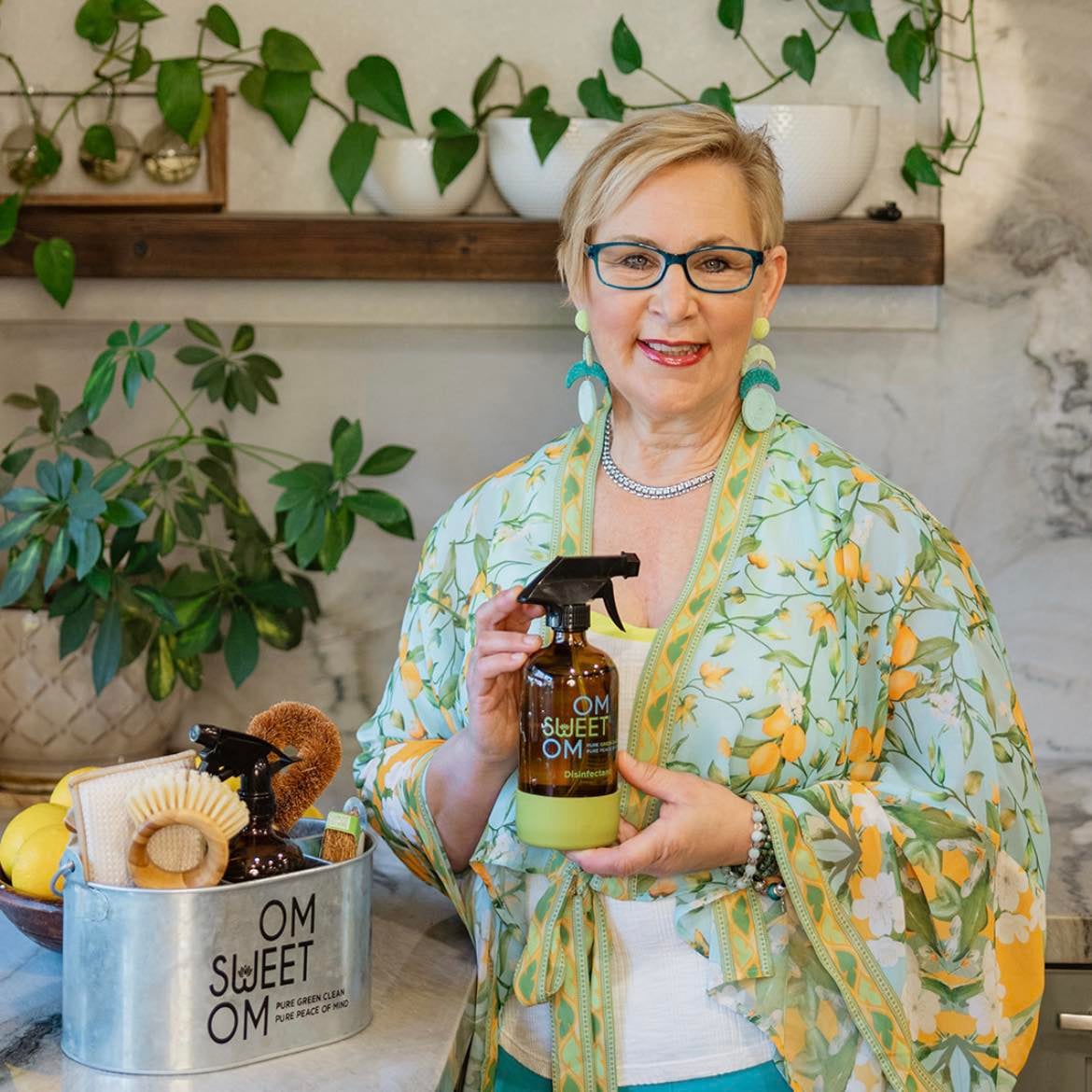 Woman holding a bottle of OM Sweet OM disinfectant in her kitchen, surrounded by eco-friendly cleaning supplies, promoting green cleaning solutions.