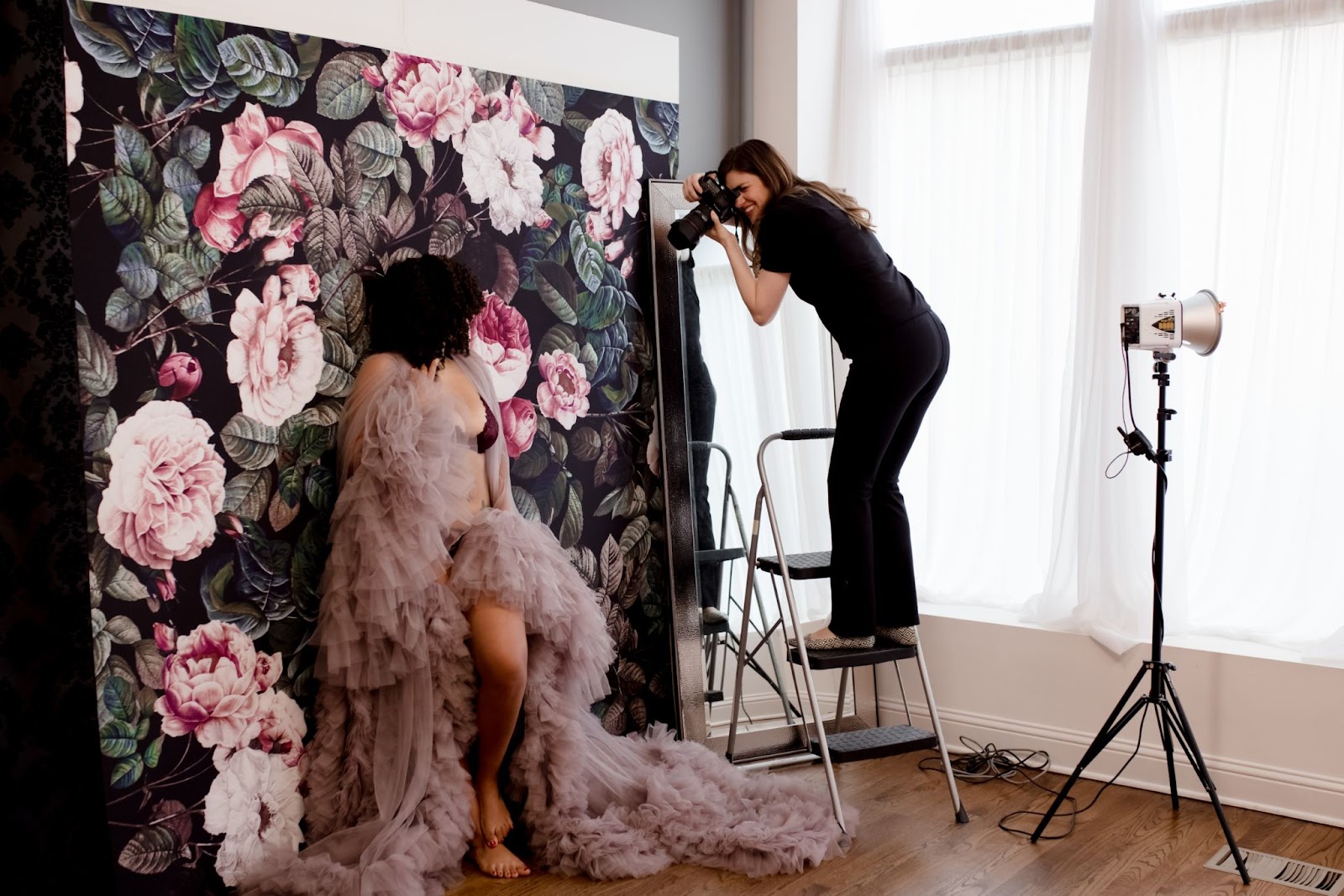 A photographer captures a boudoir portrait of a woman in a flowing tulle gown in a studio with floral backdrops.