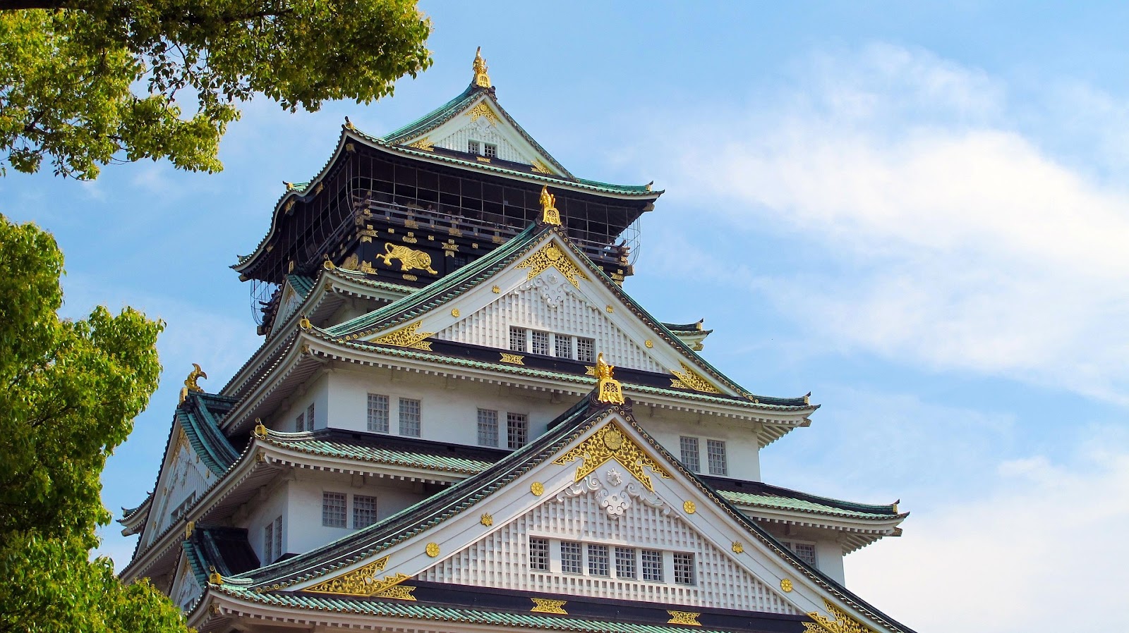 Close-up view of the Osaka Castle with traditional Japanese architecture, featuring ornate gold details and tiled roofs, surrounded by trees under a clear blue sky.