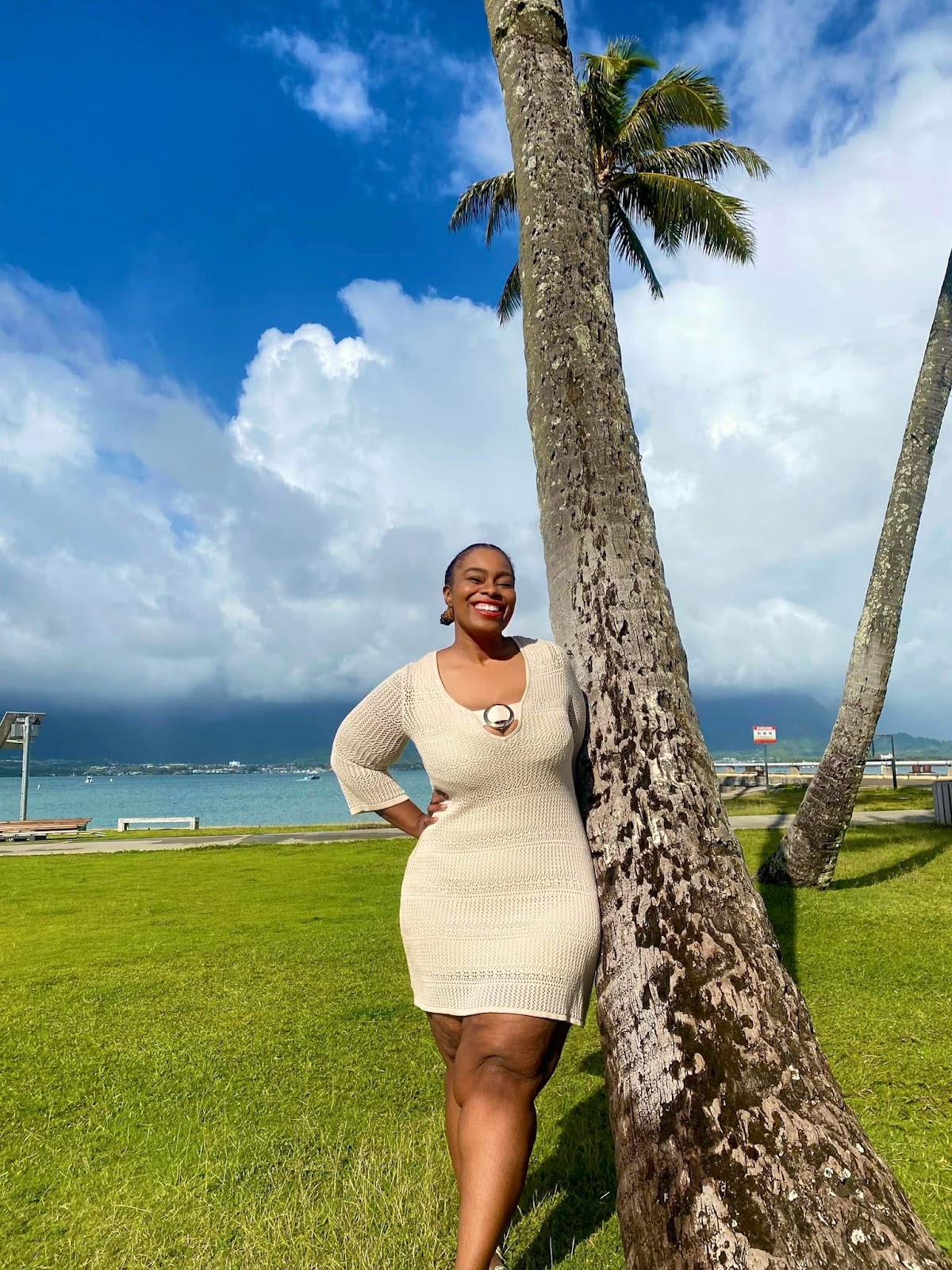 Confident woman standing by a palm tree on a sunny beach with a scenic backdrop of blue sky and ocean.