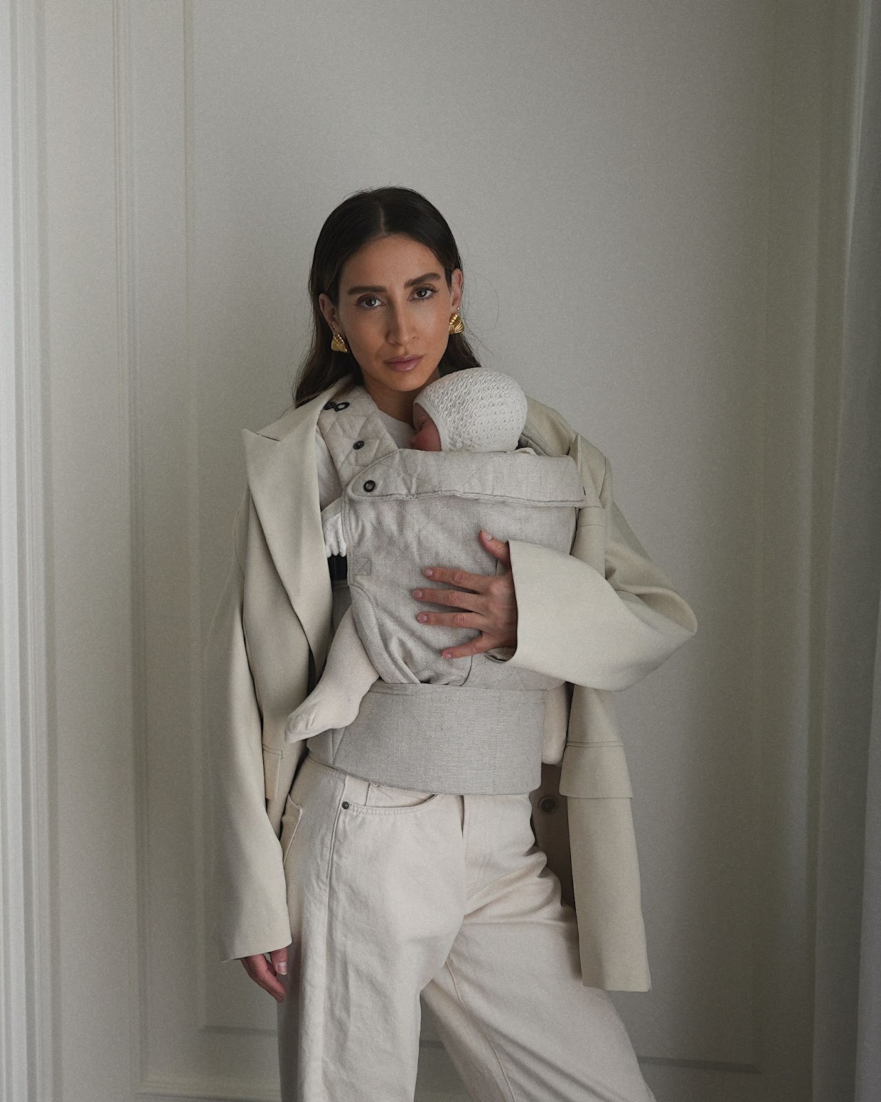 A woman holding her newborn baby in a beige baby carrier while wearing a stylish coat and beige pants, posing in a minimalist room with neutral walls and soft lighting.