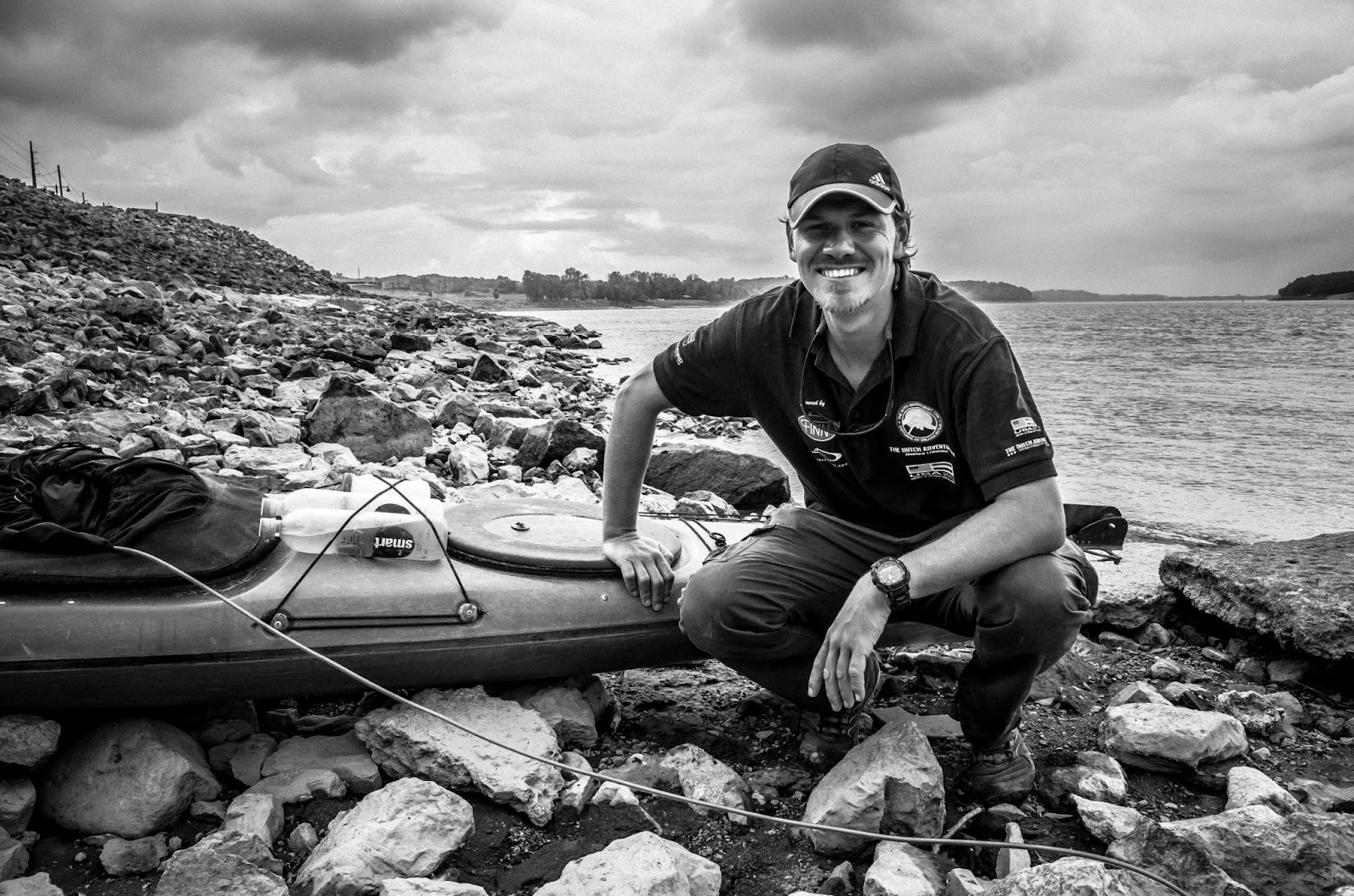 Man posing beside kayak on a rocky shore, smiling, with water and stormy skies in the background.
