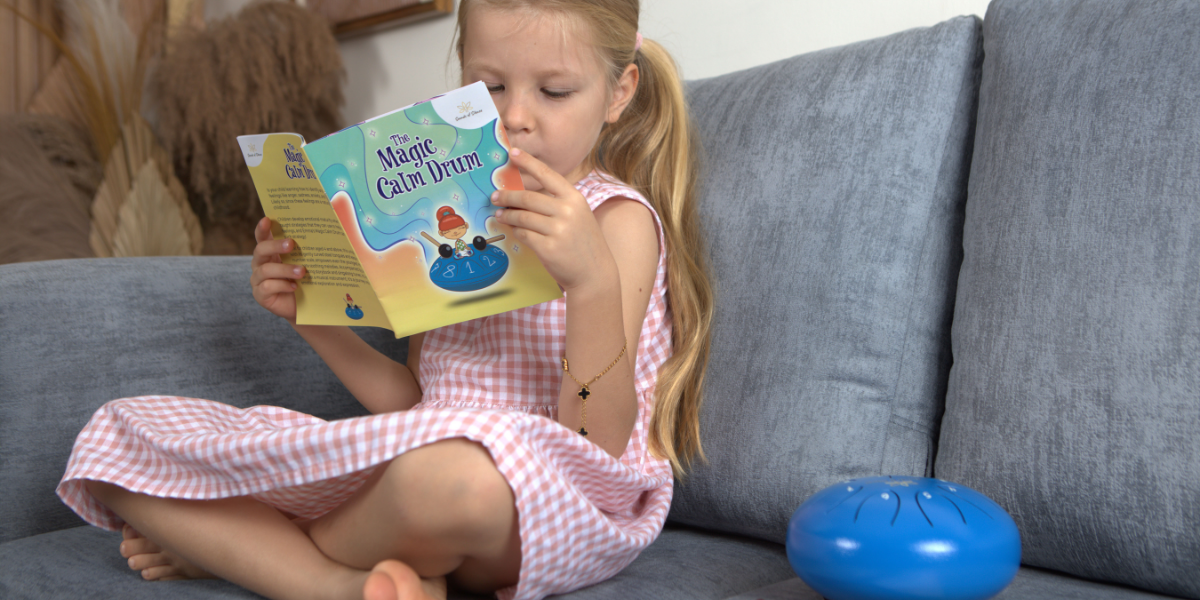 Young girl sitting on a gray couch, reading 'The Magic Calm Drum' book. Next to her is a blue handpan drum. She is dressed in a pink gingham dress and focused on the book, with a calm, relaxed expression.