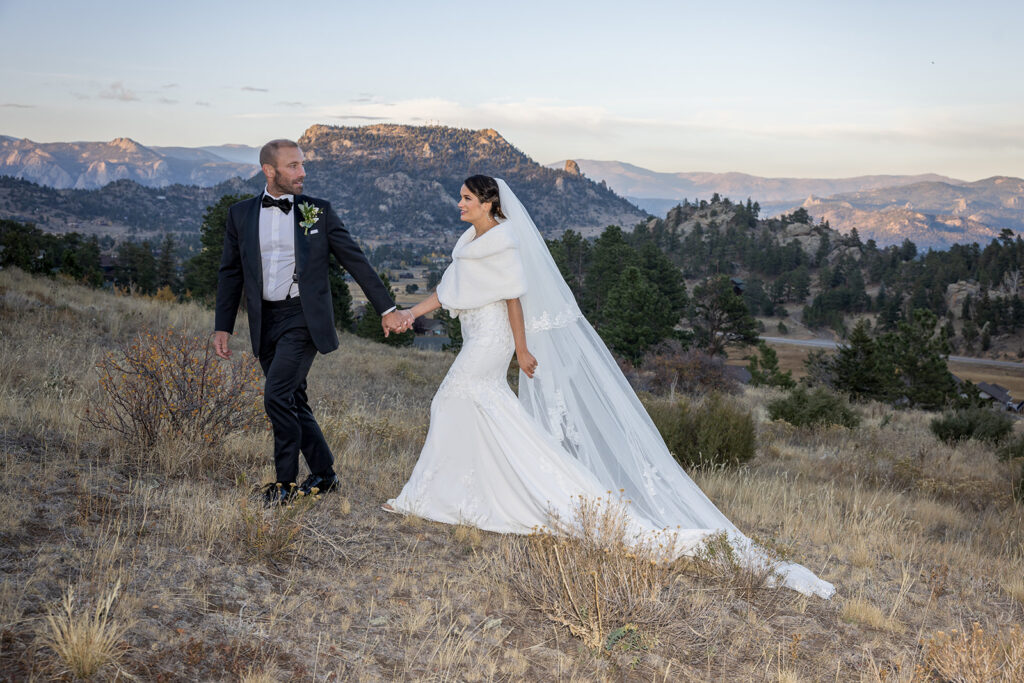 A bride and groom walking hand-in-hand in a scenic mountain setting, with the bride wearing a white gown and veil, and the groom in a tuxedo.