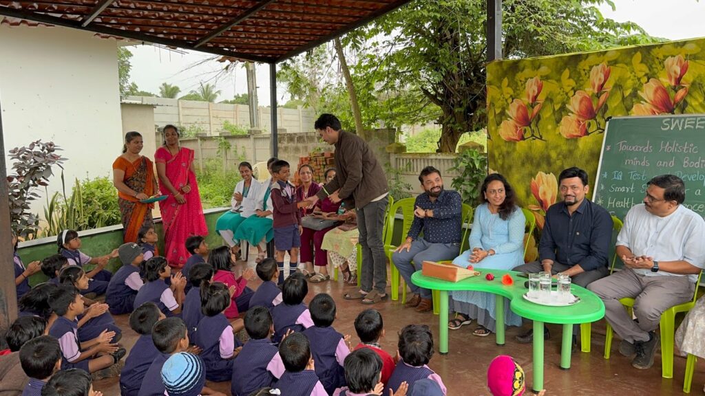 "Group of people at a school event, with children seated and a boy shaking hands with an adult, while others observe in the background. The event is focused on holistic learning and development."