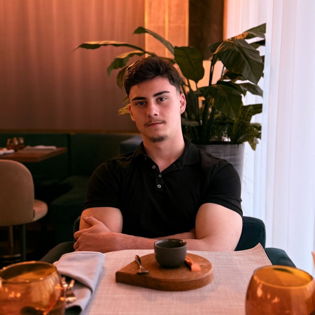 "Young man sitting at a restaurant table with a calm expression, wearing a black polo shirt, with a plant in the background and a simple dish on the table."