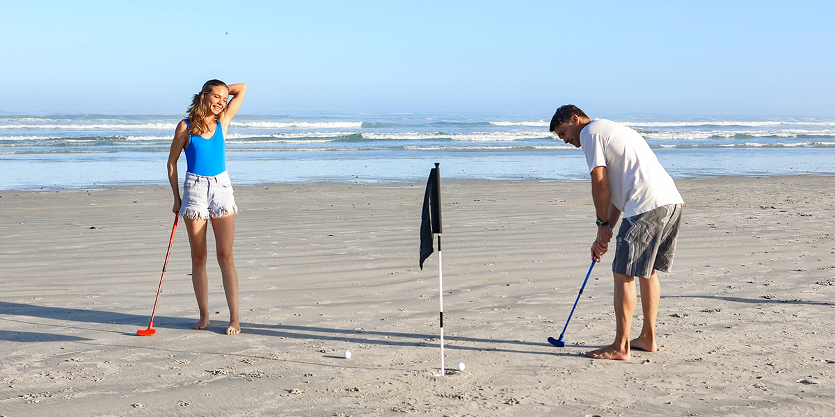 "Couple playing beach golf with colorful clubs and flags on a sunny beach with the ocean in the background."