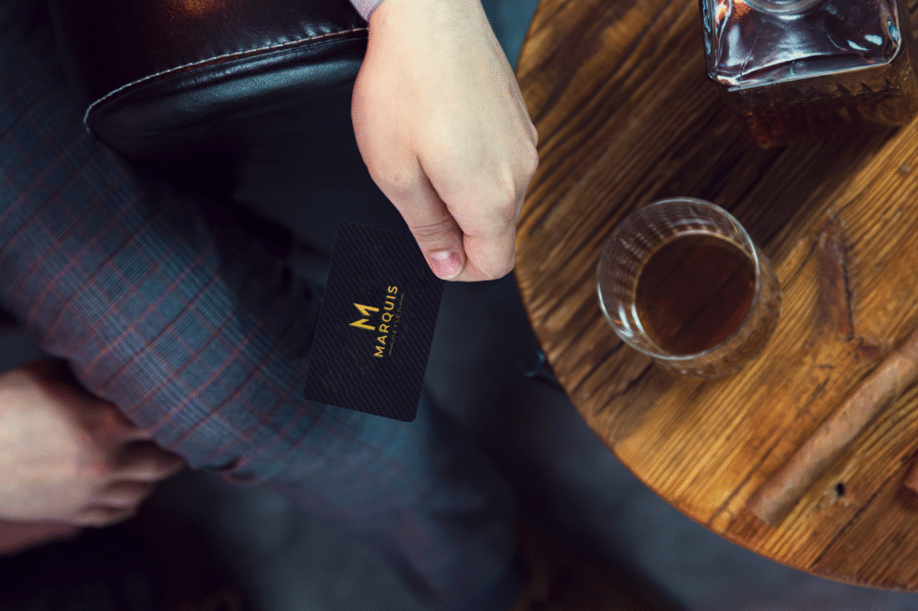 A close-up of a hand holding a sleek, black business card from Marquis, set on a wooden table with a glass of whiskey and a cigar, creating a sophisticated and elegant ambiance.