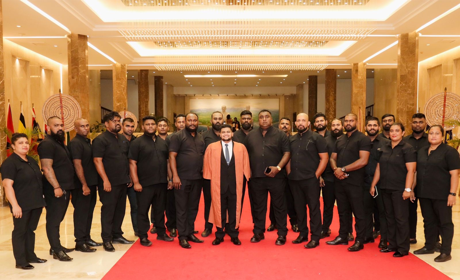Group photo of a well-dressed security team standing together with a man in a suit at the front, in a grand, luxurious setting with a red carpet.
