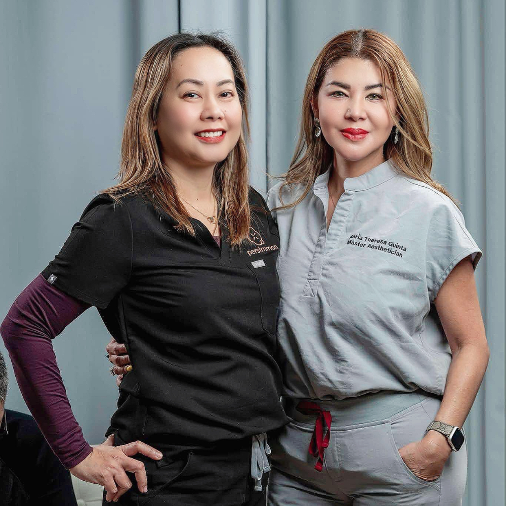 Two professional women posing in medical attire, one in a black scrub top and the other in a grey scrub top, with a soft blurred background. They are standing confidently and smiling, representing skilled professionals in the aesthetics field.