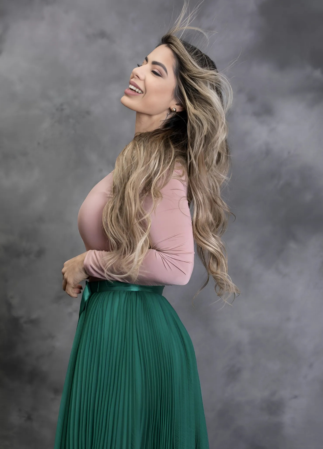 Portrait of a smiling woman in a pink long-sleeve top and green pleated skirt, with flowing hair, set against a soft, gray background.