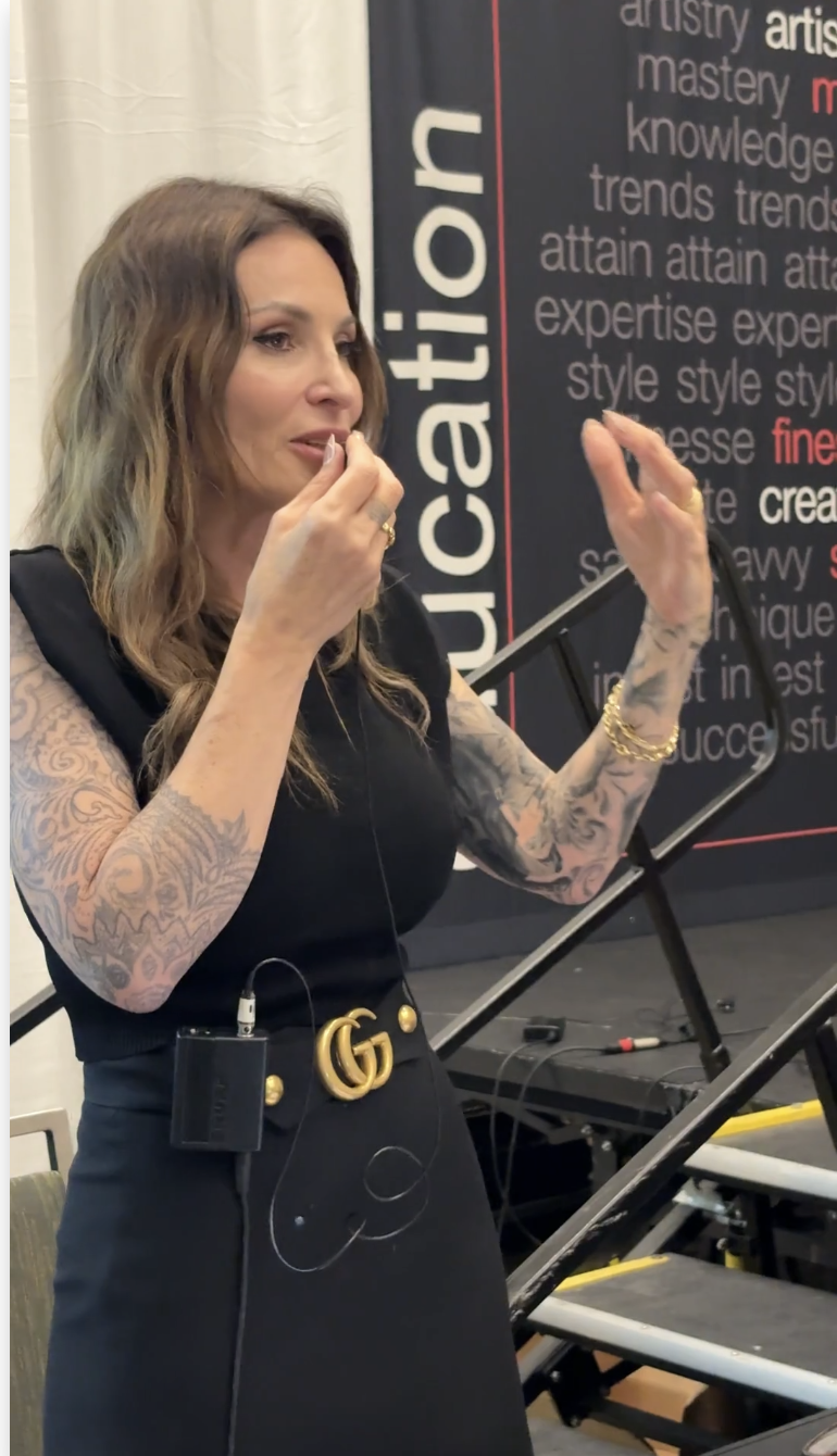 Image of a woman speaking at a seminar, gesturing with her hands while wearing a black dress and a microphone pack, standing in front of a backdrop with the word "education" and various other words related to mastery and success.
