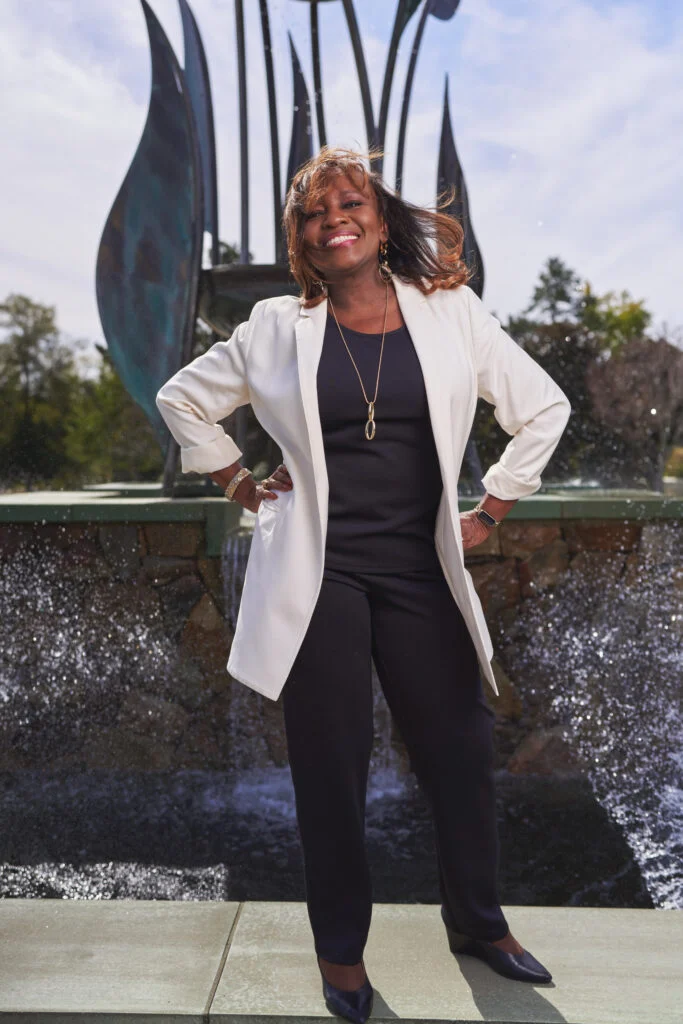 "Confident woman wearing a white jacket and black pants, posing in front of a fountain in bright daylight."
