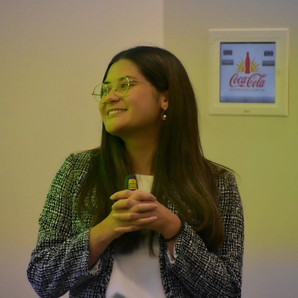 Young professional woman smiling and holding a remote control during a presentation, with a Coca-Cola advertisement visible in the background.