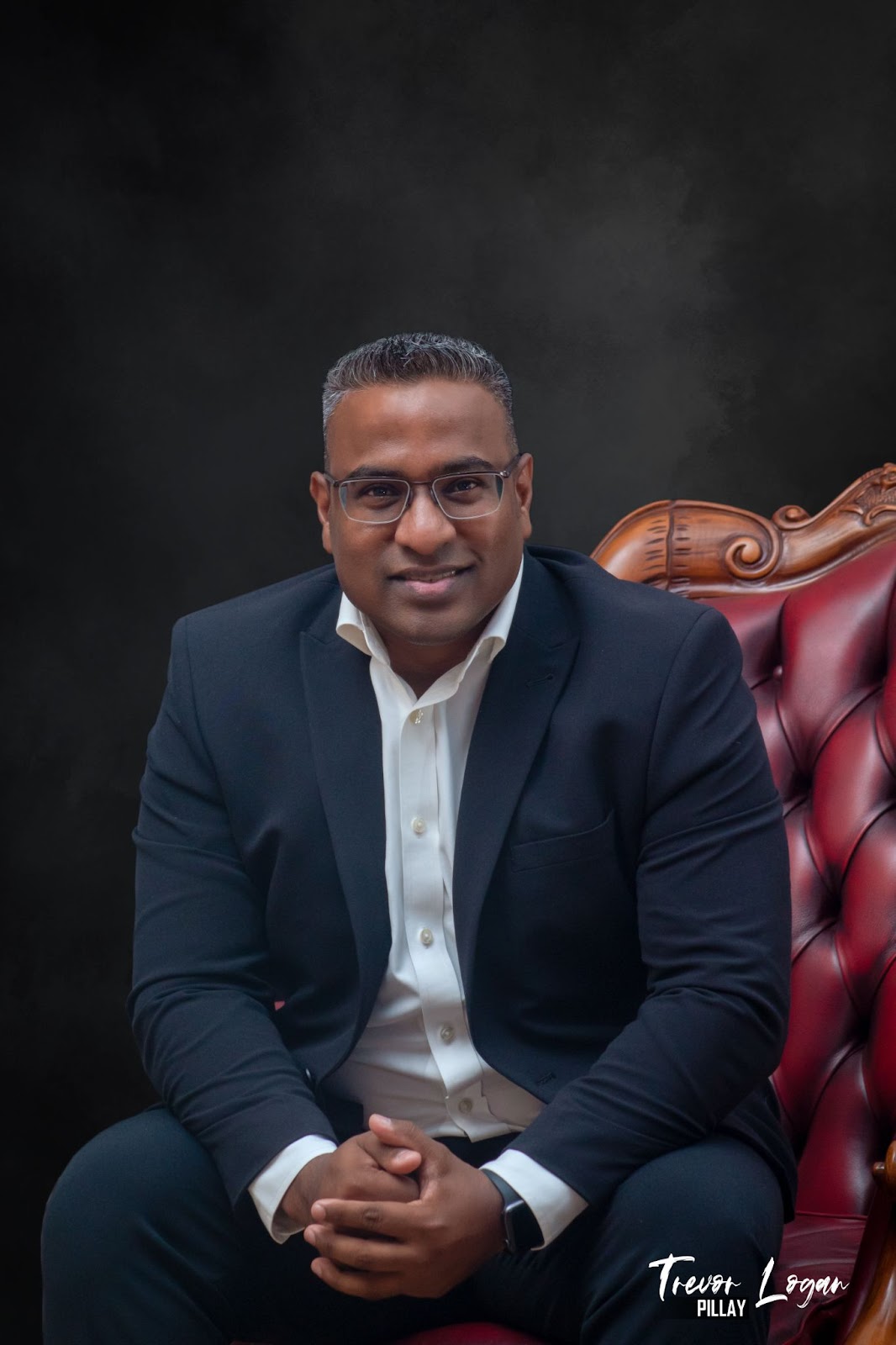 Professional portrait of a man in a black suit sitting on a red leather chair, smiling confidently with a dark background, with the name 'Trevor Logan Pillay' at the bottom."