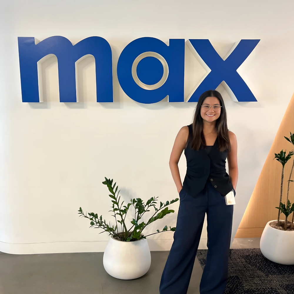 Young professional woman smiling and posing confidently in front of the 'max' company logo, standing with hands in pockets in business attire with plants in the background.