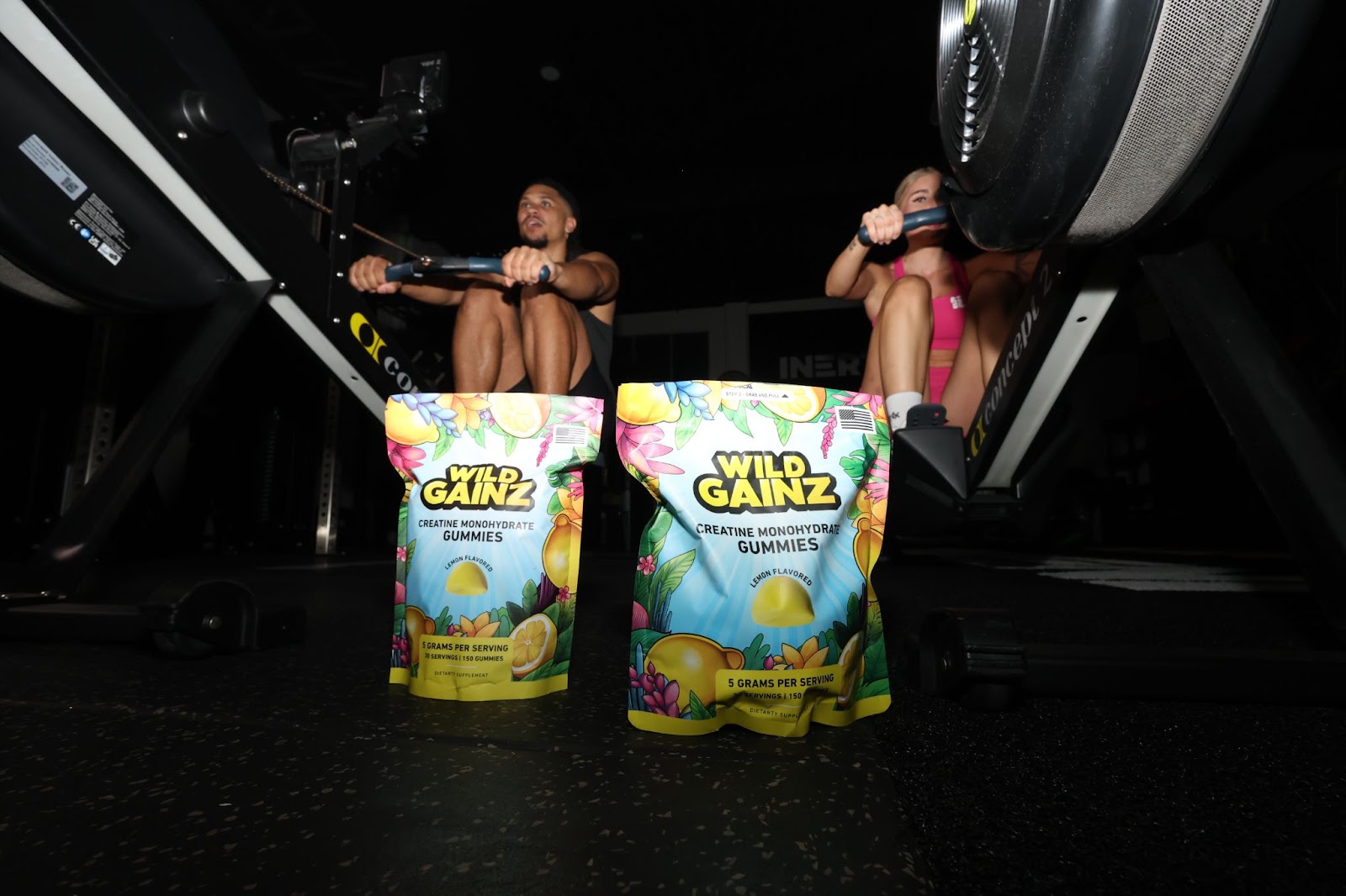 Two individuals rowing on exercise machines in a gym, with Wild Gainz Creatine Monohydrate Gummies packets placed in the foreground, highlighting the lemon-flavored supplement in a fitness setting.
