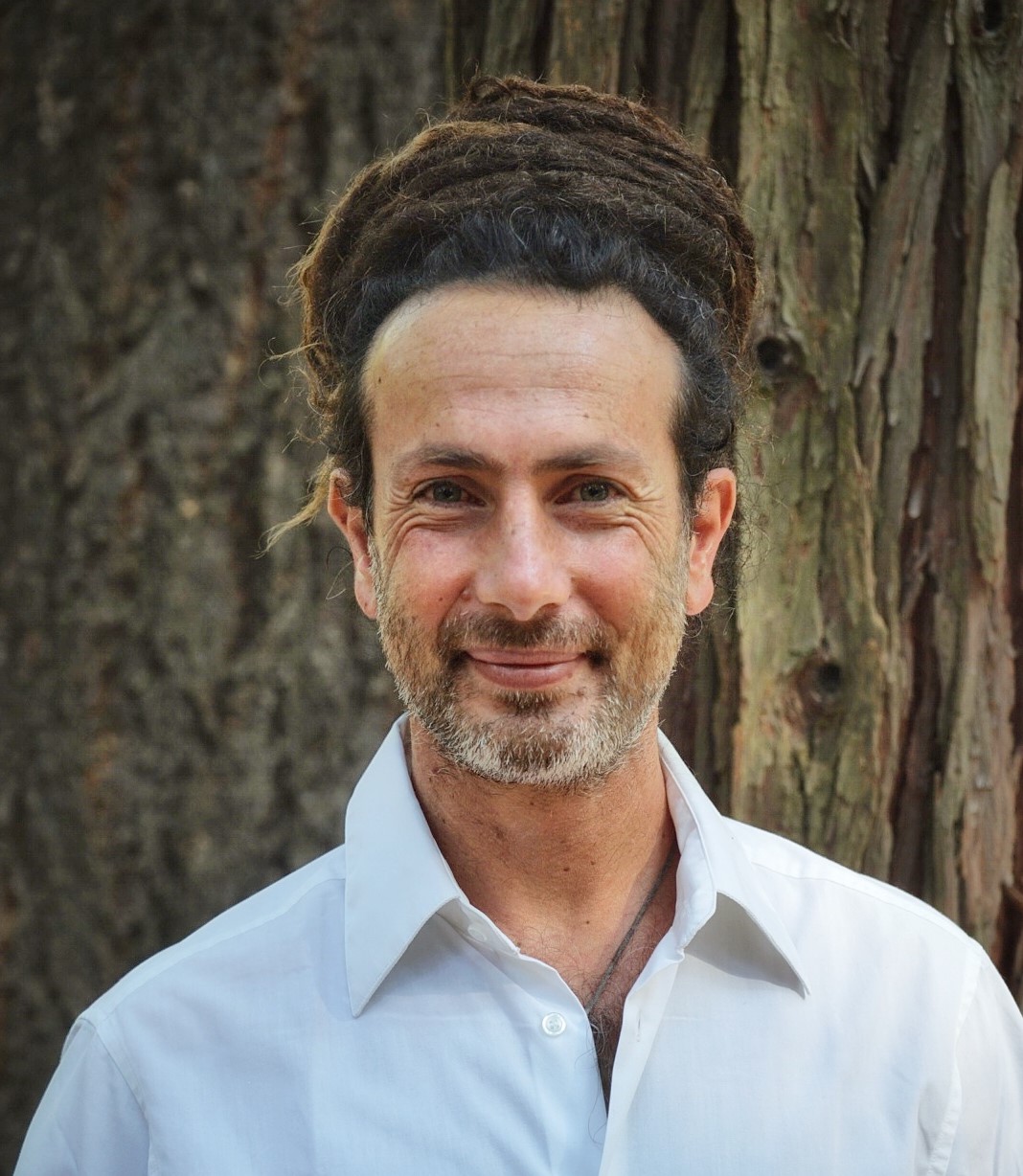 Portrait of a man with dreadlocks wearing a white shirt, smiling in front of a tree trunk, exuding a relaxed and natural vibe.