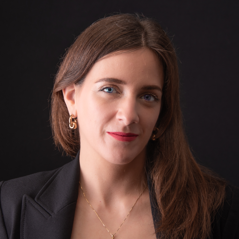 Professional woman in a black suit, with gold earrings and necklace, looking confidently at the camera against a dark background