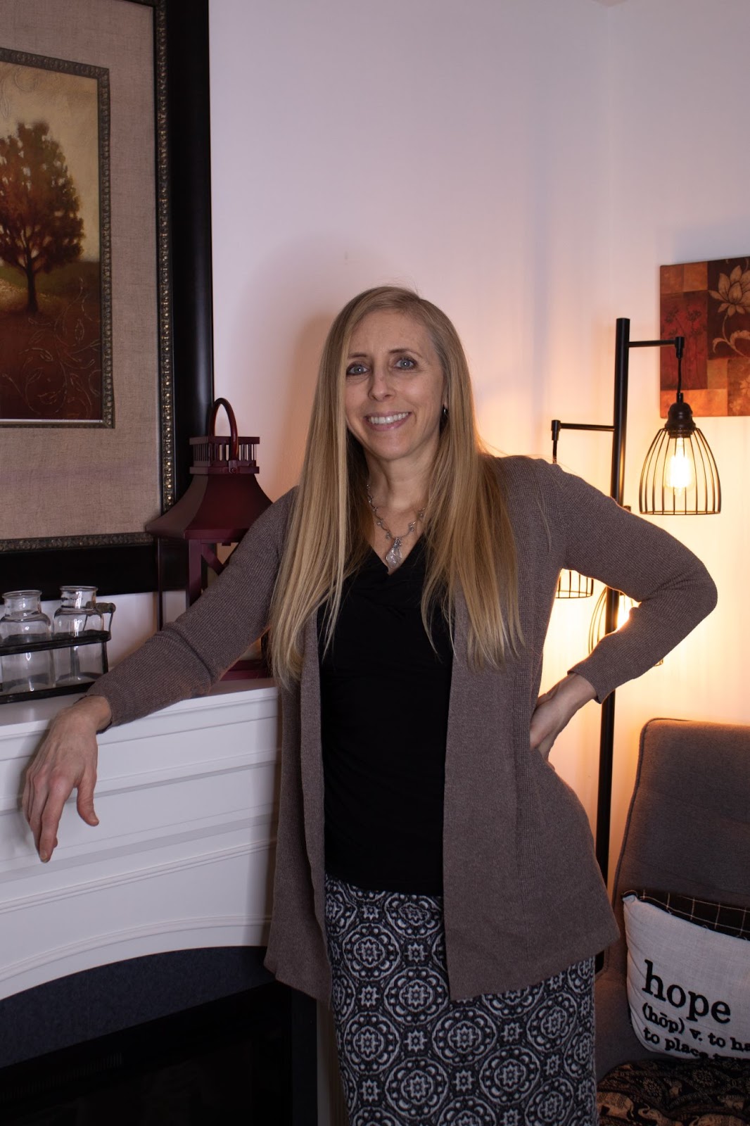 Smiling woman with long hair standing by a fireplace, wearing a brown cardigan and patterned skirt, with a cozy home setting featuring warm lighting and decorative elements in the background