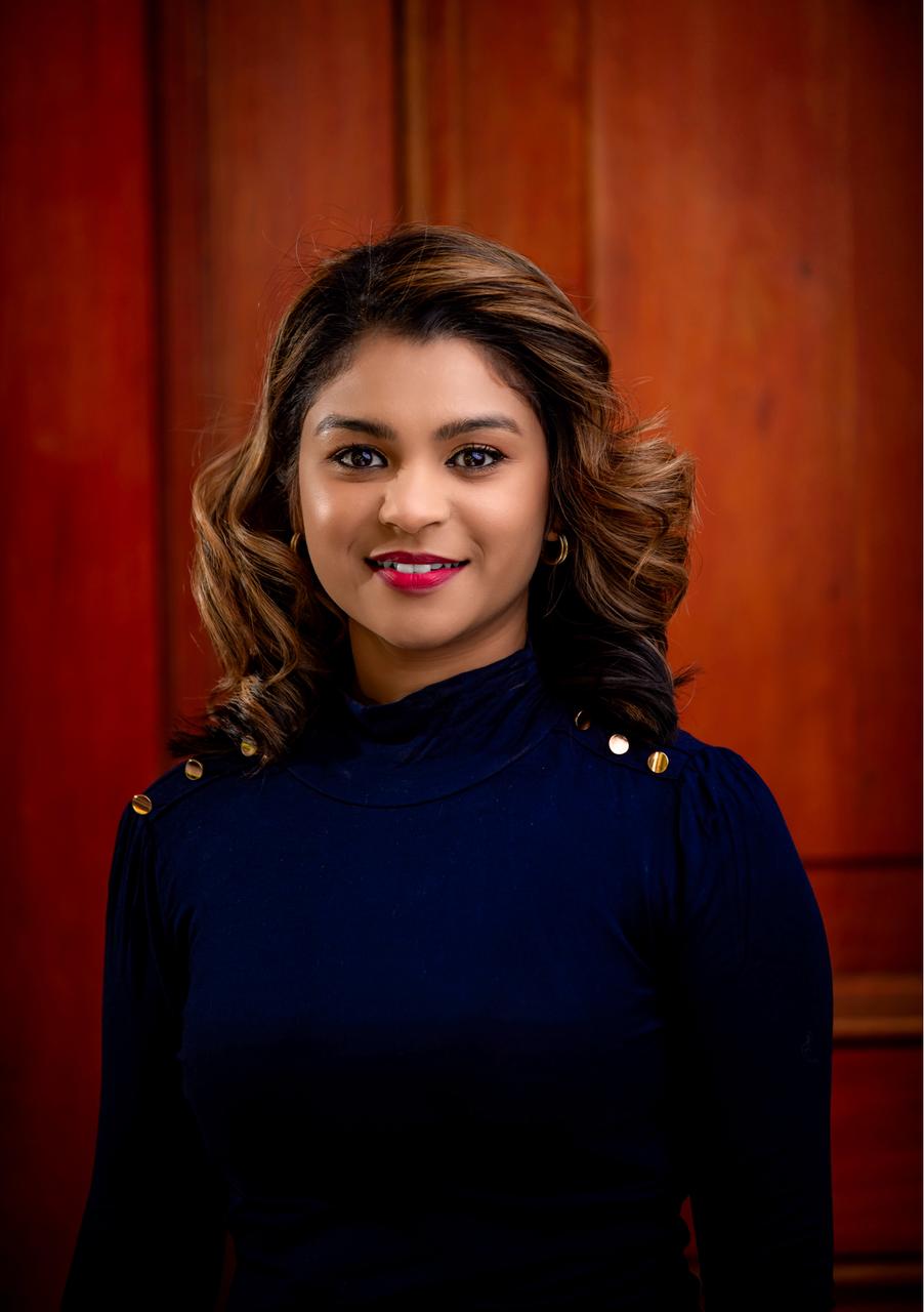 Portrait of a confident young woman in a navy blue top, with a warm smile, posing in front of a wooden backdrop.