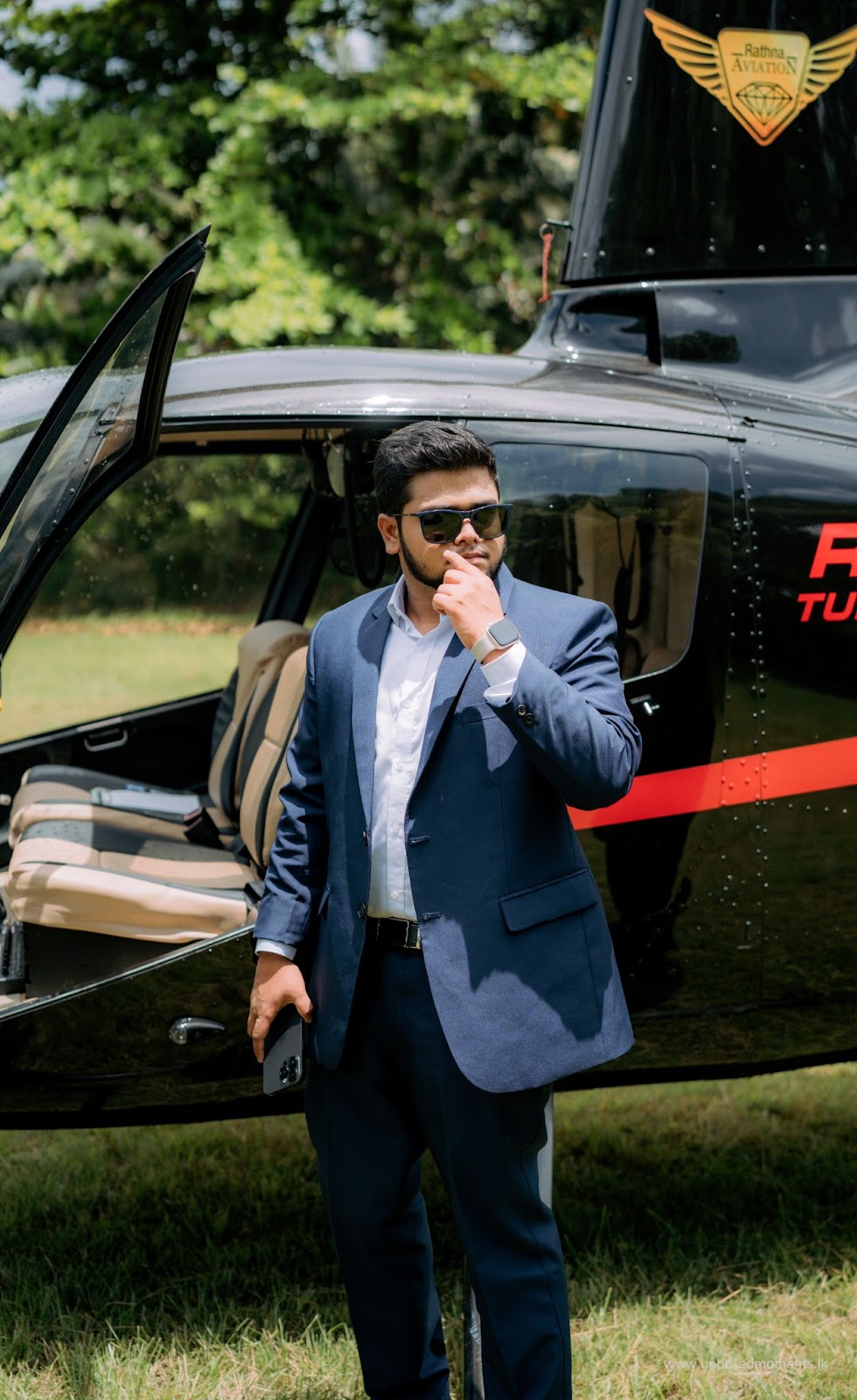 Man in a blue suit and sunglasses, standing confidently beside a helicopter in a field, with a serious and stylish demeanor