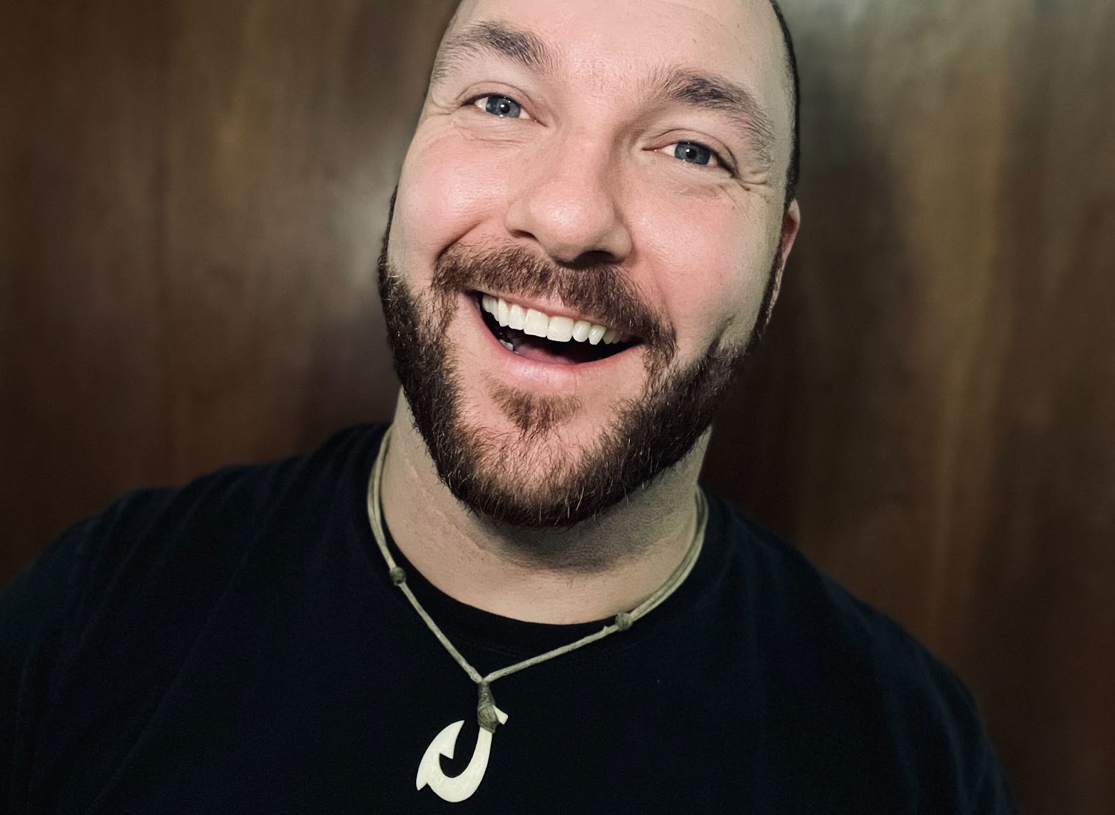 Smiling man with a beard and blue eyes wearing a black t-shirt and a necklace with a pendant, standing in front of a wooden background.