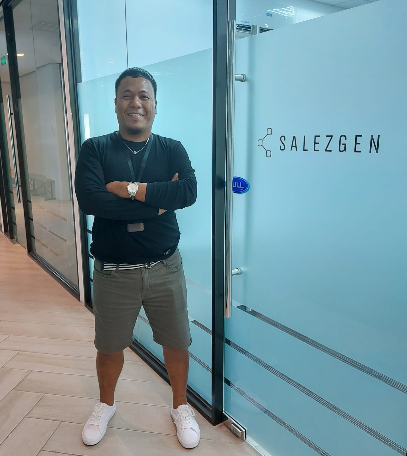 A smiling man stands with arms crossed in front of a glass office door with the 'SALEZGEN' logo, wearing a black long-sleeve shirt, gray shorts, and white sneakers, inside a modern office space.