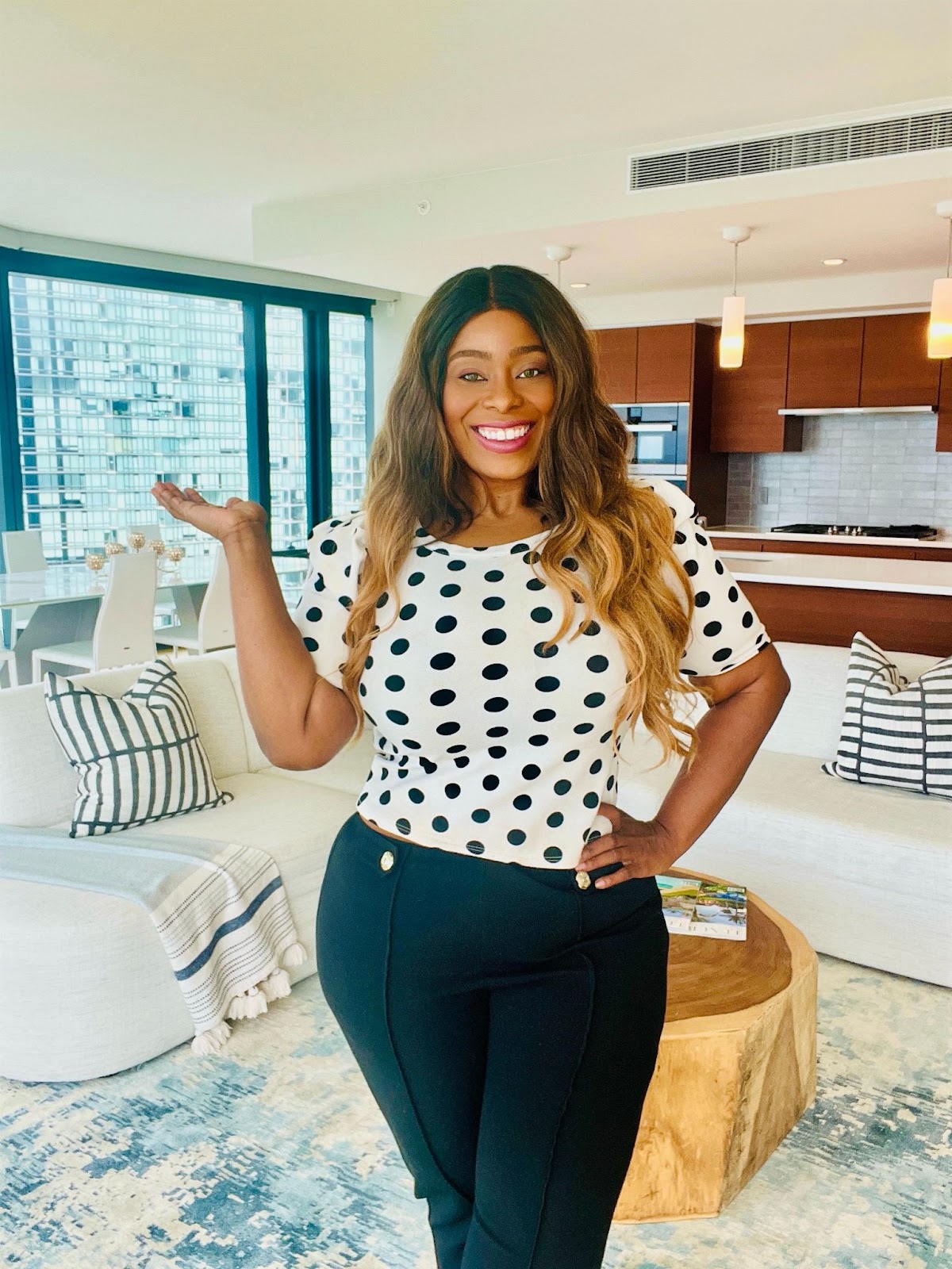 Smiling woman in a polka dot shirt and black pants posing in a modern apartment with stylish decor.