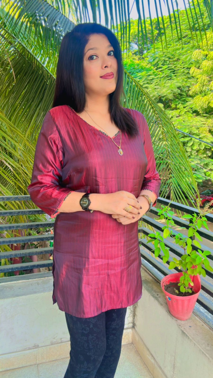 Portrait of a woman wearing a maroon tunic standing outdoors with palm trees and greenery in the background.
