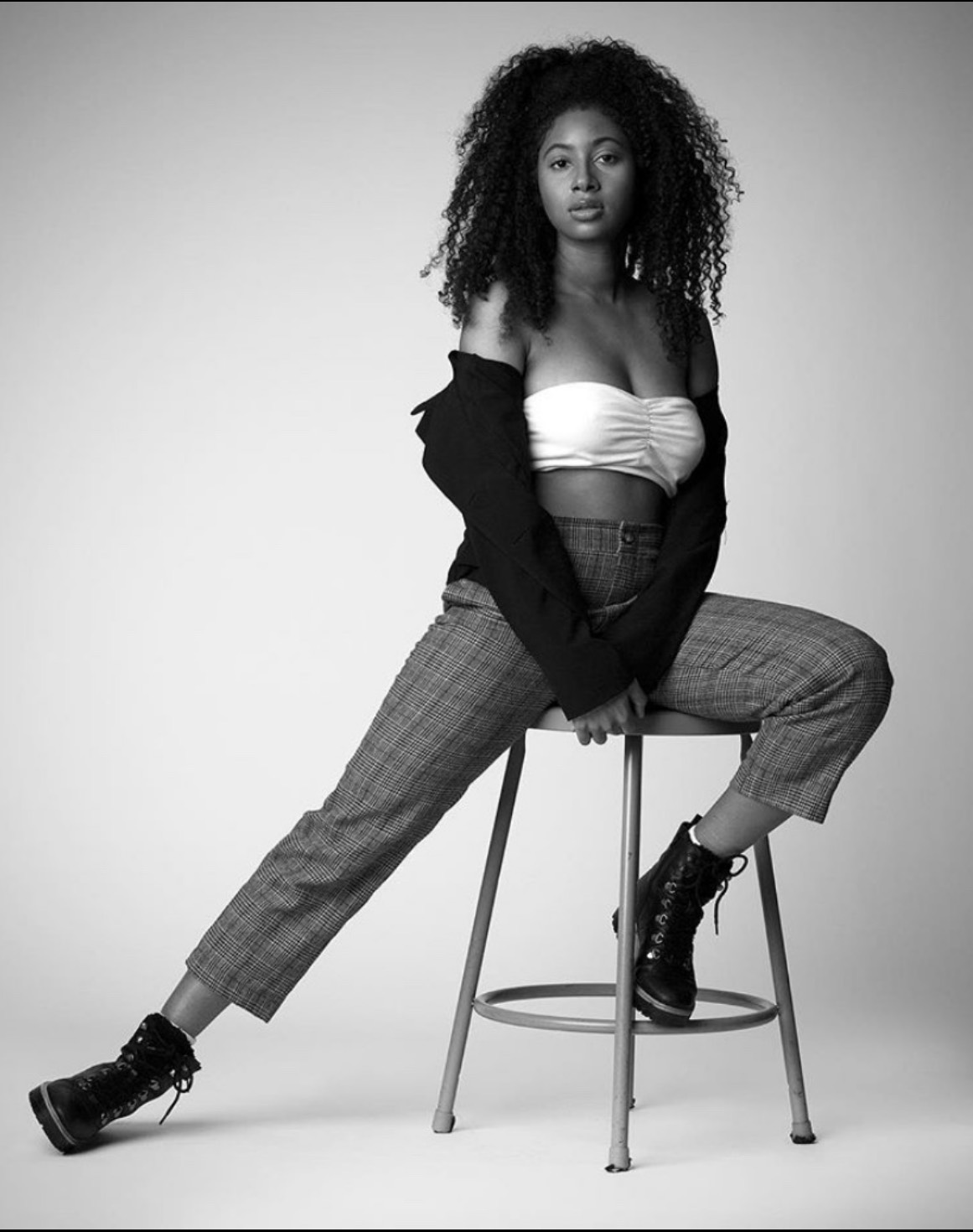 Black and white portrait of a young woman with curly hair, sitting on a stool in a relaxed pose. She is wearing a white strapless top, plaid pants, and black combat boots, with a black jacket draped over her shoulders.