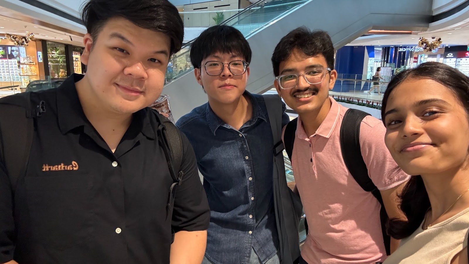 A group of four friends smiling for a selfie inside a shopping mall, featuring two men and two women wearing casual clothing and posing together