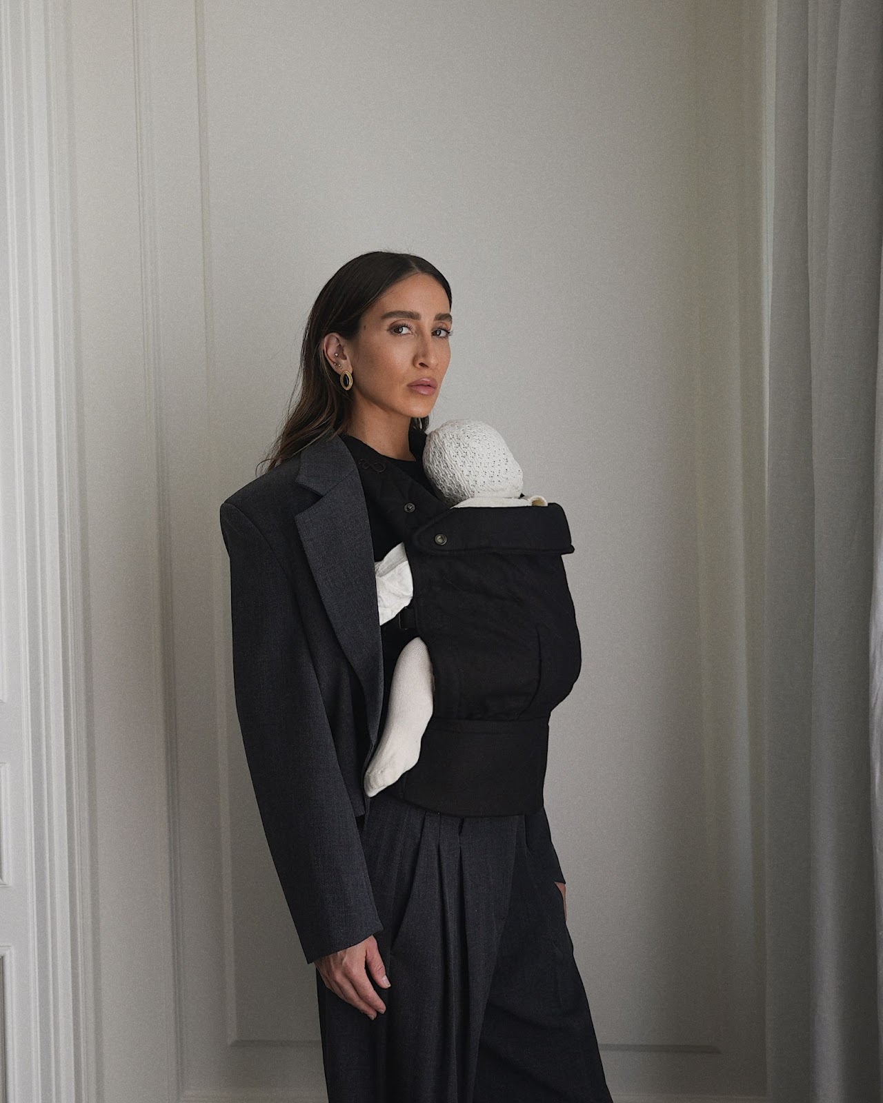 A woman carrying her baby in a black baby carrier, dressed in a black blouse and dark pants, posing in front of a light-colored wall with natural soft lighting.