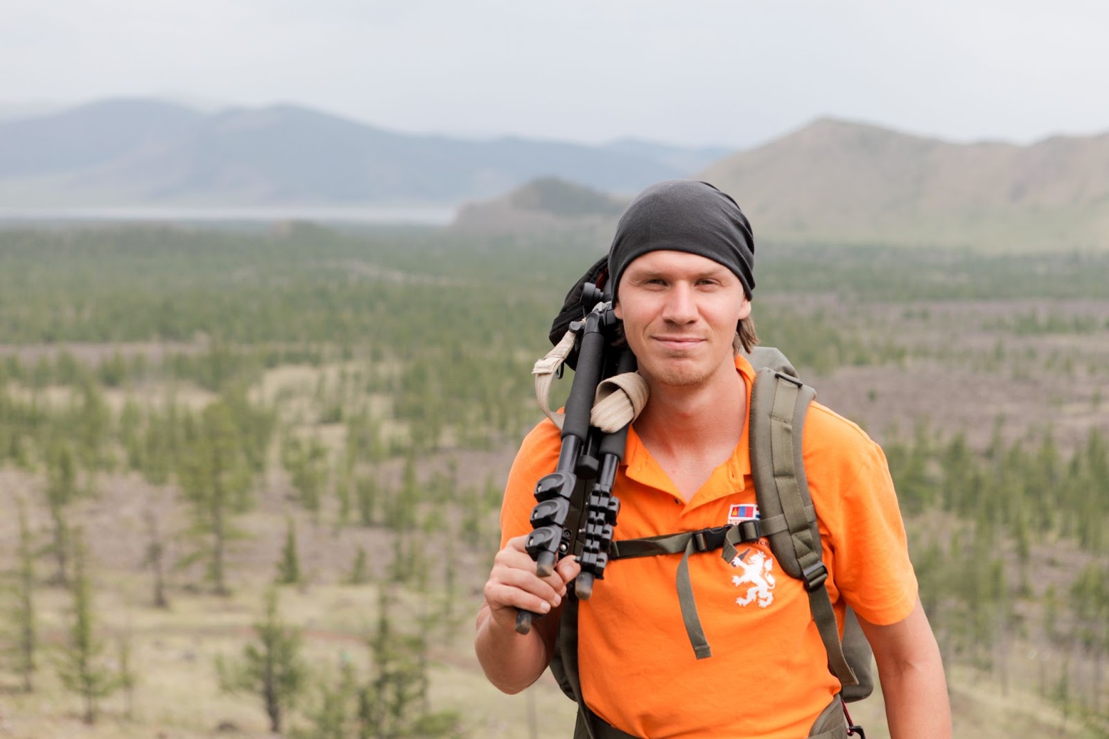 Outdoor adventurer hiking in a vast landscape with green trees and mountains, wearing a bright orange shirt and a black headband.