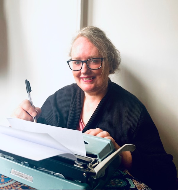 "Smiling woman sitting at a vintage typewriter, writing with a pen, showcasing her passion for writing."