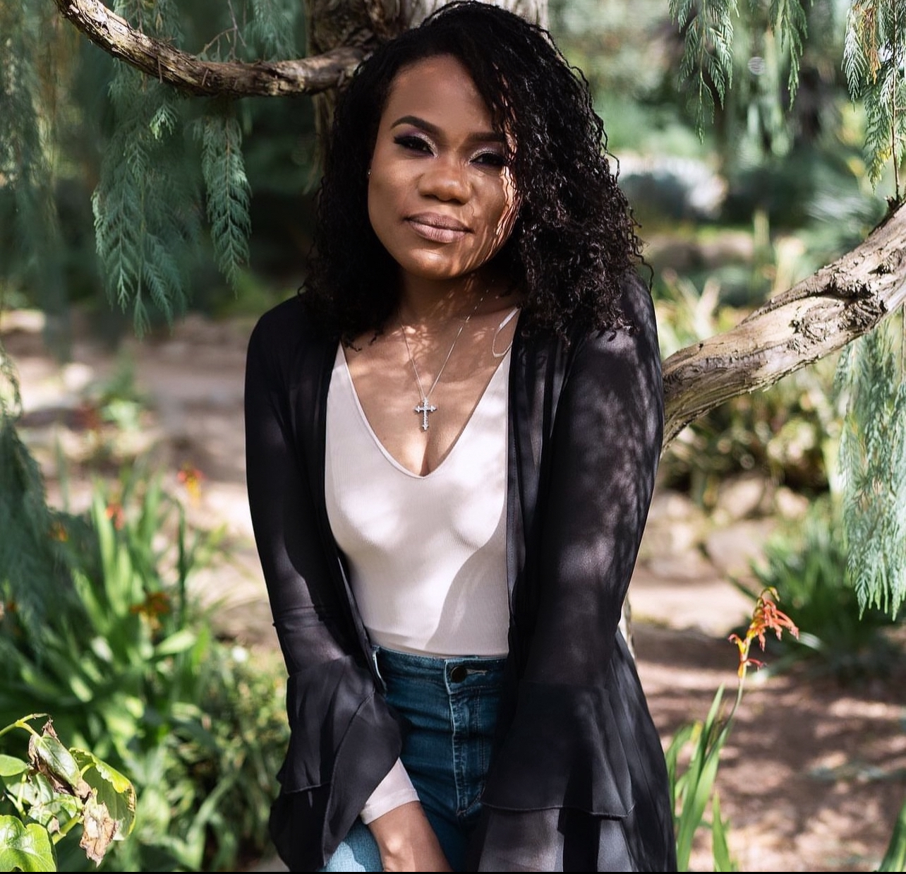 Outdoor portrait of a young woman with curly hair sitting under a tree, wearing a black jacket and light top, surrounded by greenery.