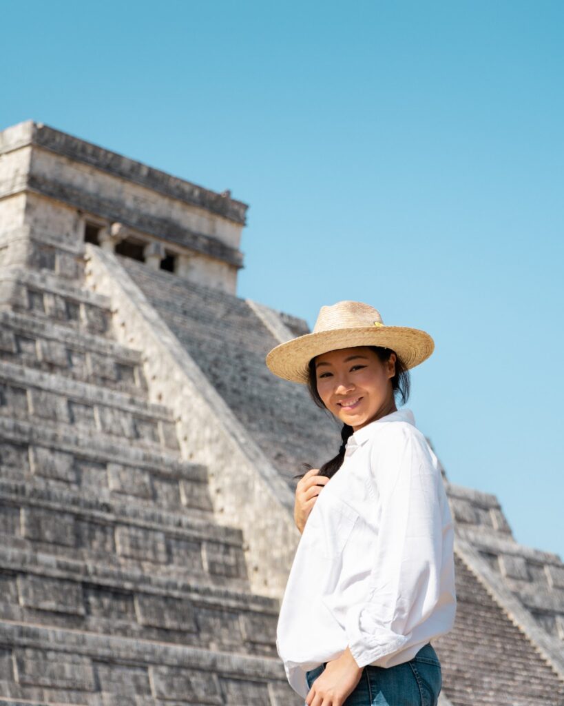 "Tourist woman in a white shirt and straw hat standing in front of the ancient Chichen Itza pyramid in Mexico, representing cultural exploration."