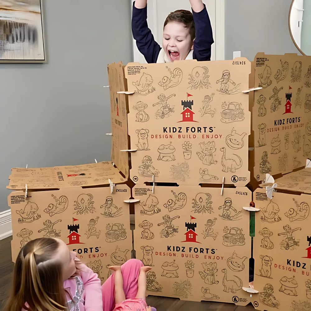 Two children enjoying playtime with Kidz Forts, a cardboard fort-building kit. One child is inside the fort, raising their arms in excitement, while the other sits on the floor nearby, surrounded by decorated cardboard pieces featuring playful illustrations