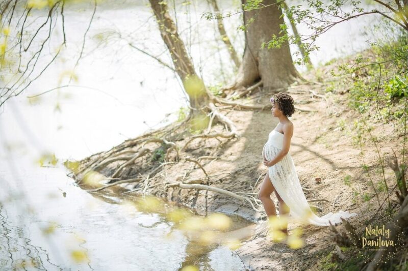 "Maternity photo of a woman standing by the water's edge, wearing a white flowing dress and a flower crown, showcasing her pregnancy in a peaceful outdoor setting."