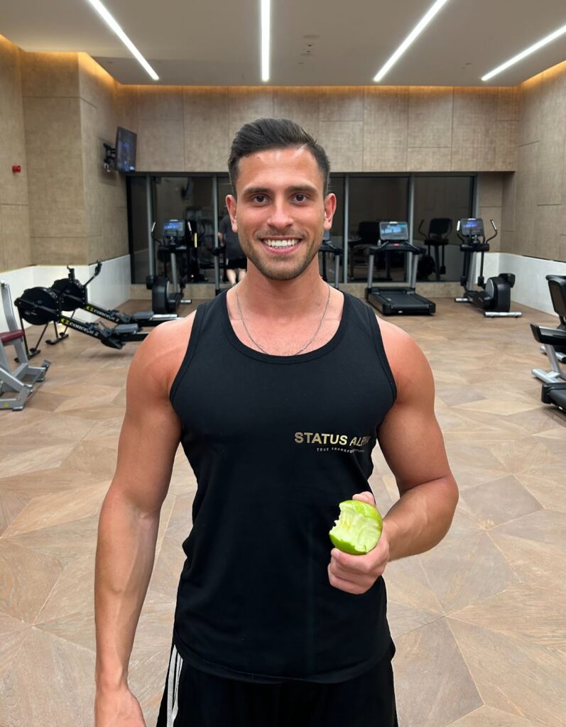 "Man in a black tank top holding a green apple, smiling at the gym with fitness equipment in the background."
