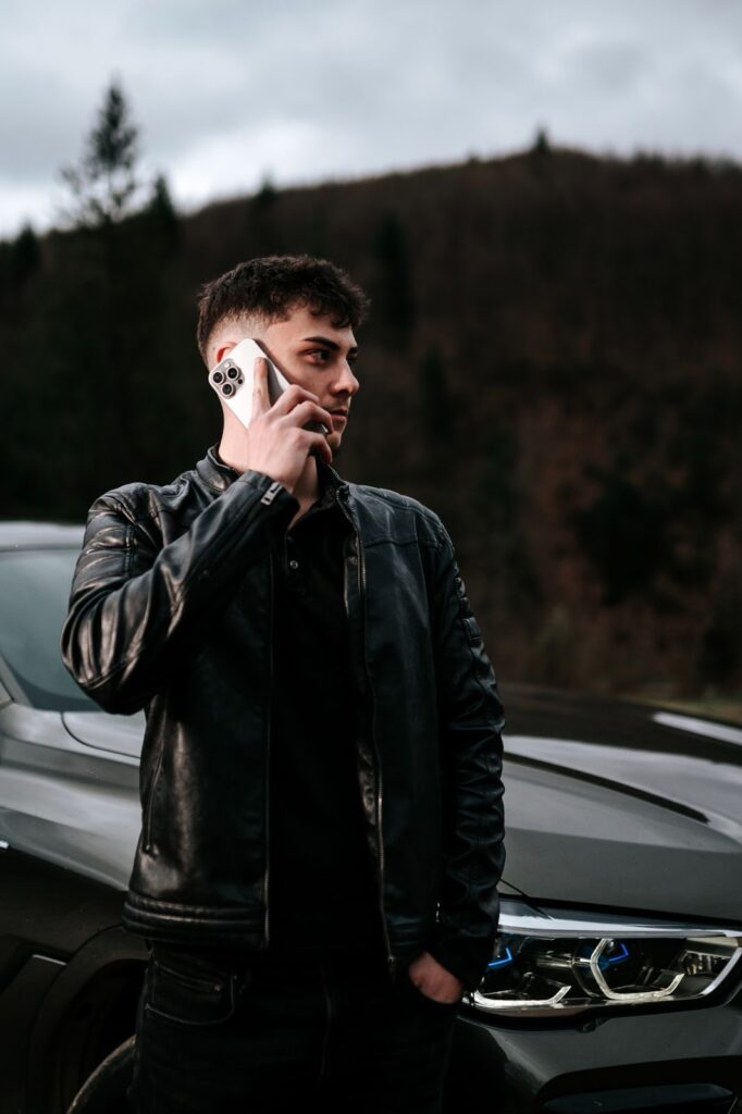 "Young man in a black leather jacket talking on the phone next to a sleek car, surrounded by nature with a dramatic sky in the background."